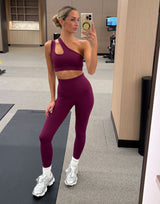 A woman takes a mirror selfie in a gym while wearing a matching burgundy activewear set consisting of a one-shoulder sports bra and high-waisted leggings, paired with white socks and silver sneakers.