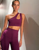 A woman modeling a matching athletic set consisting of a one-shoulder crop top and high-waisted leggings in a deep plum purple color, set against a textured beige wall.