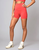 A model wearing high-waisted coral pink athletic biker shorts, a matching sports bra, and white sneakers, standing against a neutral gray studio background.