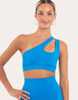 A woman modeling a bright blue one-shoulder crop top with a side cutout, paired with matching high-waisted bottoms, against a plain light beige studio background.