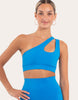 A woman modeling a bright blue one-shoulder crop top with a side cutout, paired with matching high-waisted bottoms, against a plain light beige studio background.