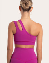 Rear view of a woman wearing a bright magenta one-shoulder sports bra and matching high-waisted leggings against a light beige studio background.