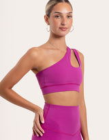 A woman modeling a bright magenta one-shoulder sports bra and matching high-waisted leggings against a light beige studio background.