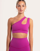 A woman modeling a bright magenta one-shoulder sports bra and matching high-waisted leggings against a plain light beige studio background.