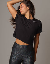 A woman modeling a black short-sleeve crop top with a small white script logo on the chest, paired with dark iridescent leggings, against a solid gray studio background.