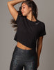 A woman modeling a black short-sleeve crop top with a small white script logo on the chest, paired with dark iridescent leggings, against a solid gray studio background.