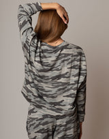 Person wearing a camouflage outfit with one arm raised on a plain background